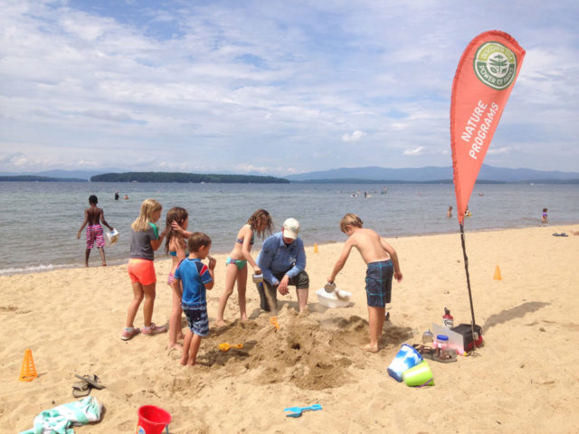 A ranger and a group of kids at a beach. They are working together to build large sandcastles that the ranger will in turn use to show how lakes are formed.