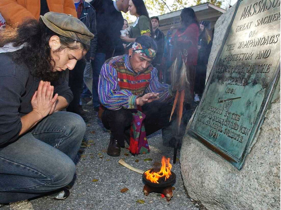 Two people kneel over a small fire in front of a historic marker.