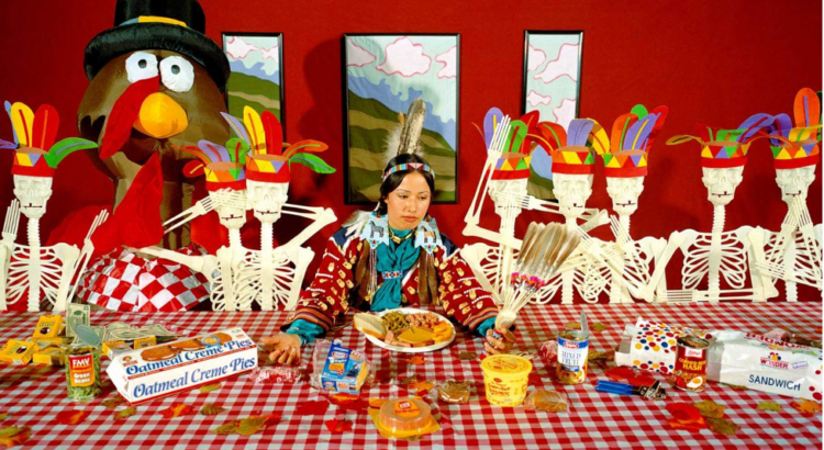 A woman wearing a feathered headdress sits at a red table surrounded by plastic skeletons in headdresses.