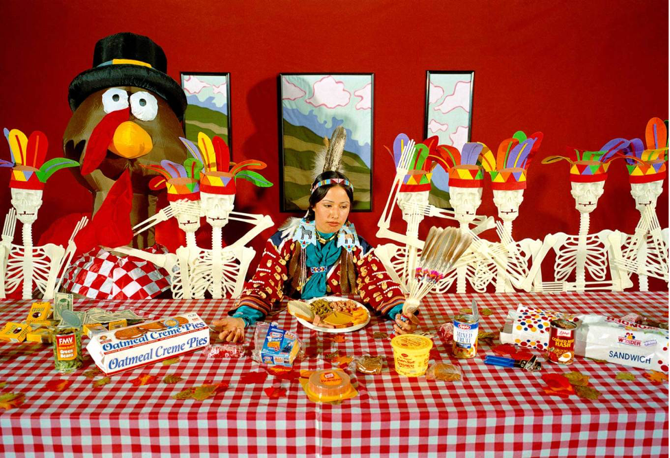 A woman wearing a feathered headdress sits at a red table surrounded by plastic skeletons in headdresses