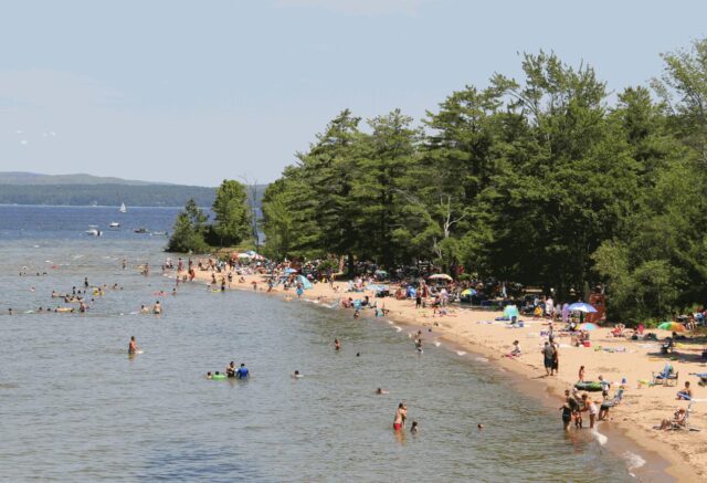 A sandy beach next to a lake with people swimming and sun bathing. 